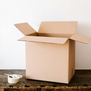Opened cardboard container placed on old wooden desk near tape and scissors before relocation against white wall
